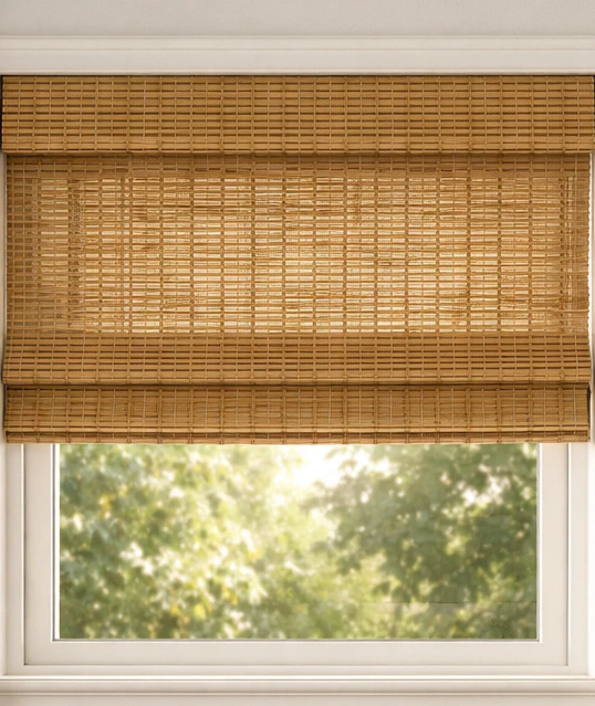 Close-up of a natural woven bamboo roman shade mounted inside a white window frame, half-raised showing the bamboo slat texture and warm afternoon light streaming through trees behind the window glass below