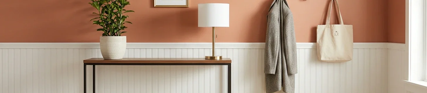 Close-up of a styled entryway corner showing the two-material transition zone — warm terra-cotta matte painted accent wall on the upper half meeting white-painted beadboard wainscot at chair-rail height on the lower half, narrow walnut console table with a green leafy plant in a ceramic pot, brass-finish modern table lamp, partial view of a small framed botanical print, grey wool coat and canvas tote bag hanging on matte black wall hooks on the right