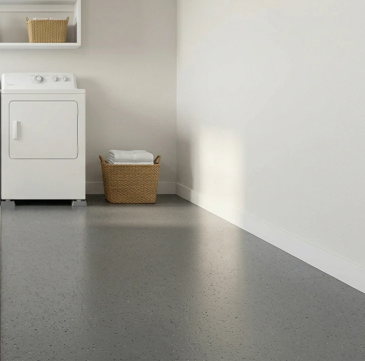 Compact laundry room corner with smooth charcoal-grey poured epoxy concrete flooring with subtle decorative speckle flake pattern catching the light, white top-load-style dryer on the left edge, woven wicker basket of folded white towels in the corner, partial view of a white floating utility shelf with a small basket top-left and clean white walls, showing the seamless industrial-utility surface that prevents washer-leak penetration with zero seam path through to the slab