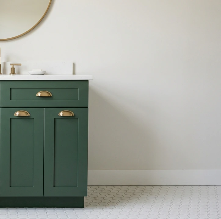 Bathroom vanity painted matte forest green with brushed brass cup-pull hardware, white quartz countertop, brass faucet and small soap dish, round brass mirror partially visible at top and white hex tile floor below, showing the bonded paint finish that handles bathroom humidity