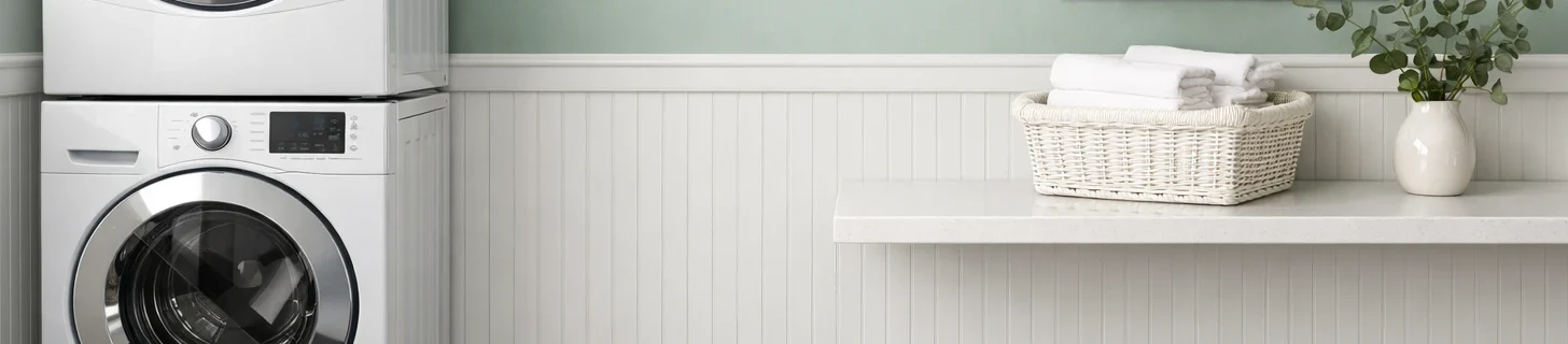 Close-up of a laundry room two-material wall treatment showing the transition zone — soft sage-green matte painted upper wall meeting white-painted PVC beadboard wainscot at chair-rail height on the lower half, partial view of a stacked white front-load washer-dryer on the left, white quartz folding counter on the right holding a white wicker basket of folded towels and a small ceramic vase with eucalyptus branches