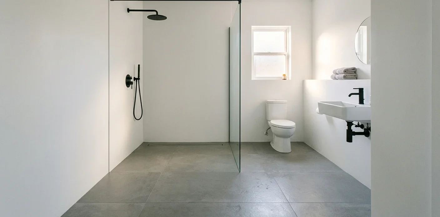 Small curbless wet room bathroom with a continuous large-format grey porcelain floor sloping to a slim linear drain along the shower wall, a single fixed frameless glass panel, matte black rain shower and hand shower, white walls, a small frosted window, a wall-hung white basin with black tap and folded towels above it